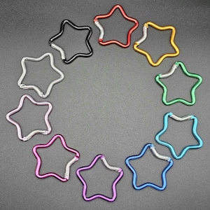 Puede incluir: Una colecci&oacute;n de diez mosquetones con forma de estrella dispuestos en c&iacute;rculo. Cada mosquet&oacute;n presenta un color met&aacute;lico diferente, incluyendo rojo, dorado, verde, azul claro, azul oscuro, morado, magenta, rosa, plateado y negro, con un cierre de resorte plateado.
