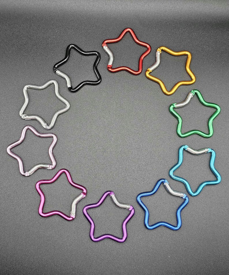 Puede incluir: Una colecci&oacute;n de diez mosquetones con forma de estrella dispuestos en c&iacute;rculo. Cada mosquet&oacute;n presenta un color met&aacute;lico diferente, incluyendo rojo, dorado, verde, azul claro, azul oscuro, morado, magenta, rosa, plateado y negro, con un cierre de resorte plateado.
