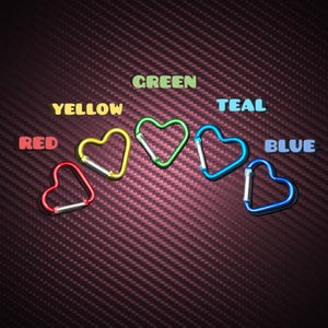 May include: Five heart-shaped carabiners in red, yellow, green, teal, and blue, each featuring a spring-loaded gate. The text labels RED, YELLOW, GREEN, TEAL, and BLUE are positioned above their respective colored carabiners on a dark background.