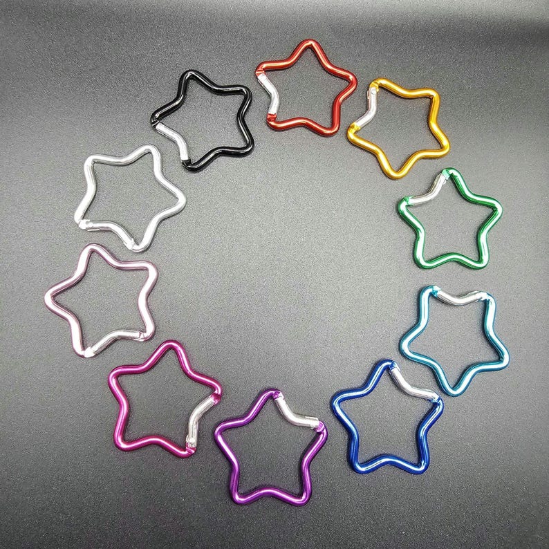Puede incluir: Un conjunto de diez mosquetones con forma de estrella dispuestos en un patr&oacute;n circular. Cada mosquet&oacute;n cuenta con un cierre de resorte y viene en un color met&aacute;lico diferente, incluyendo rojo, dorado, verde, azul claro, azul oscuro, morado, rosa, plateado y negro.