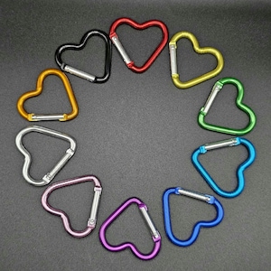 May include: A collection of ten heart-shaped carabiners arranged in a circle. The carabiners feature a metallic finish in various colors including red, yellow, green, blue, purple, pink, silver, orange, and black, each with a spring-loaded gate.