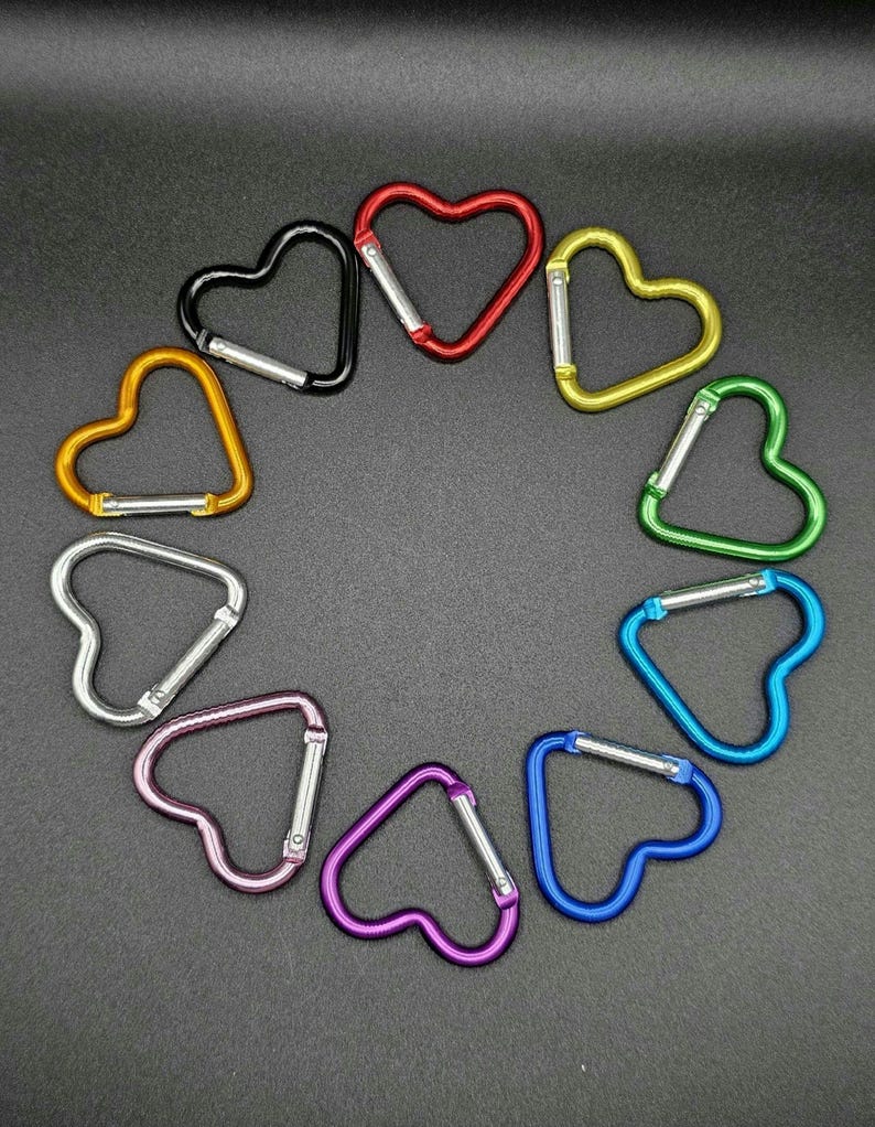 May include: A collection of ten heart-shaped carabiners arranged in a circle. The carabiners feature a metallic finish in various colors including red, yellow, green, blue, purple, pink, silver, orange, and black, each with a spring-loaded gate.