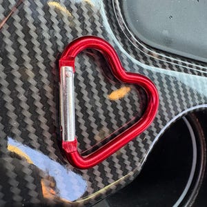 May include: A heart-shaped carabiner with a metallic red finish and a silver-colored spring-loaded gate mechanism. The carabiner features a smooth, rounded frame that forms a distinct heart silhouette, designed for securing items with a clip.