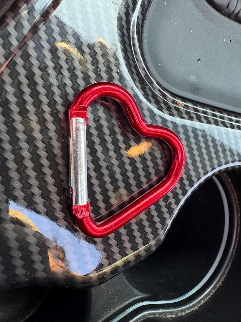 May include: A heart-shaped carabiner with a metallic red finish and a silver-colored spring-loaded gate mechanism. The carabiner features a smooth, rounded frame that forms a distinct heart silhouette, designed for securing items with a clip.