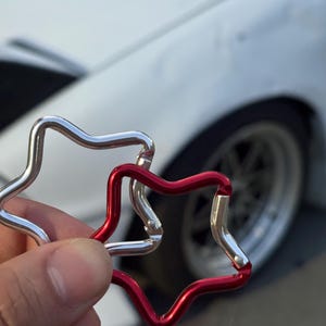 Puede incluir: Dos mosquetones en forma de estrella, uno con acabado plateado met&aacute;lico y otro con acabado rojo vibrante. Cada mosquet&oacute;n cuenta con un mecanismo de cierre con resorte que permite abrir y cerrar la estructura de estrella de metal liso.