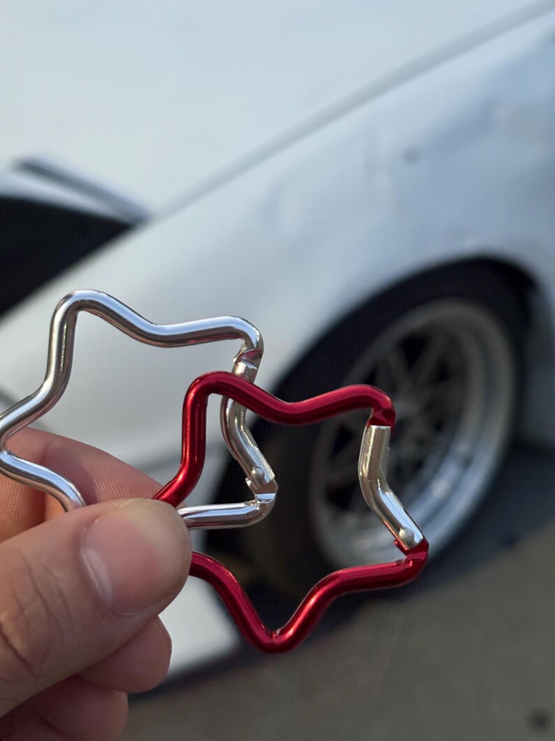 Puede incluir: Dos mosquetones en forma de estrella, uno con acabado plateado met&aacute;lico y otro con acabado rojo vibrante. Cada mosquet&oacute;n cuenta con un mecanismo de cierre con resorte que permite abrir y cerrar la estructura de estrella de metal liso.
