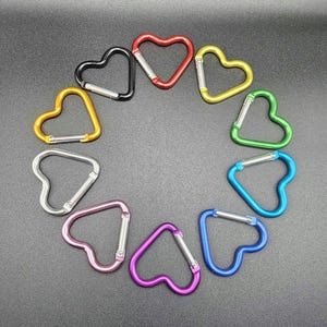 May include: A collection of ten heart-shaped carabiners arranged in a circle. The carabiners are colored red, yellow, green, light blue, dark blue, purple, pink, silver, orange, and black, each featuring a spring-loaded silver metal gate.