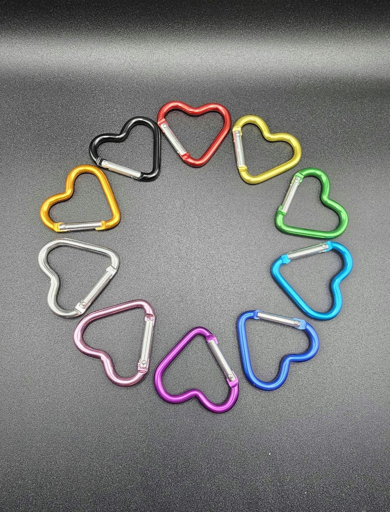 May include: A collection of ten heart-shaped carabiners arranged in a circle. The carabiners are colored red, yellow, green, light blue, dark blue, purple, pink, silver, orange, and black, each featuring a spring-loaded silver metal gate.