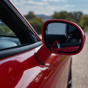 May include: A red car side mirror featuring a small, red, heart-shaped carabiner attached to the lower corner of the glass. The mirror surface displays the standard safety text, OBJECTS IN MIRROR ARE CLOSER THAN THEY APPEAR, in red lettering.