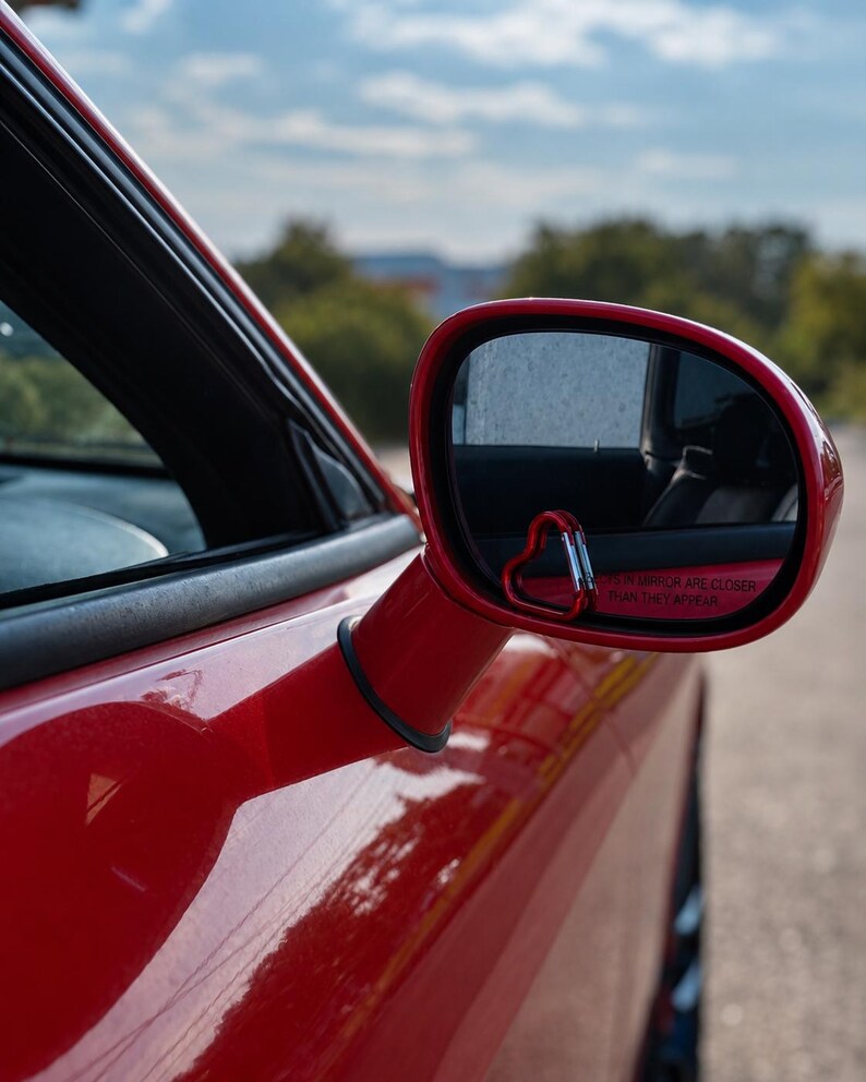 May include: A red car side mirror featuring a small, red, heart-shaped carabiner attached to the lower corner of the glass. The mirror surface displays the standard safety text, OBJECTS IN MIRROR ARE CLOSER THAN THEY APPEAR, in red lettering.