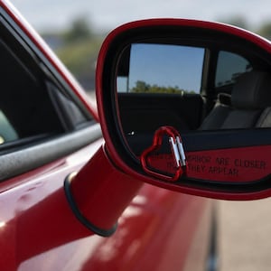 May include: A red heart-shaped carabiner attached to the lower corner of a red car side mirror. The mirror glass features the text OBJECTS IN MIRROR ARE CLOSER THAN THEY APPEAR, reflecting the interior of the vehicle and the outdoor environment.