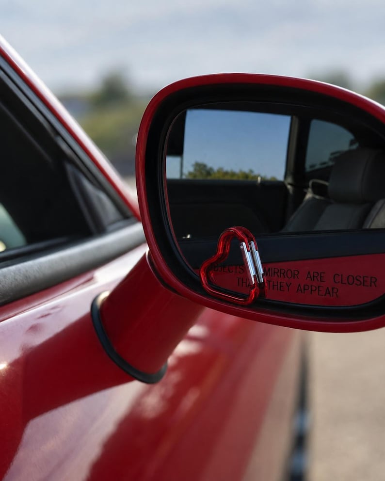 May include: A red heart-shaped carabiner attached to the lower corner of a red car side mirror. The mirror glass features the text OBJECTS IN MIRROR ARE CLOSER THAN THEY APPEAR, reflecting the interior of the vehicle and the outdoor environment.