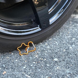 May include: Two gold-colored metal carabiners, one shaped like a five-pointed star and the other in a standard D-shape, featuring silver-toned spring-loaded gates, positioned near the base of a black rubber tire with a glossy rim.