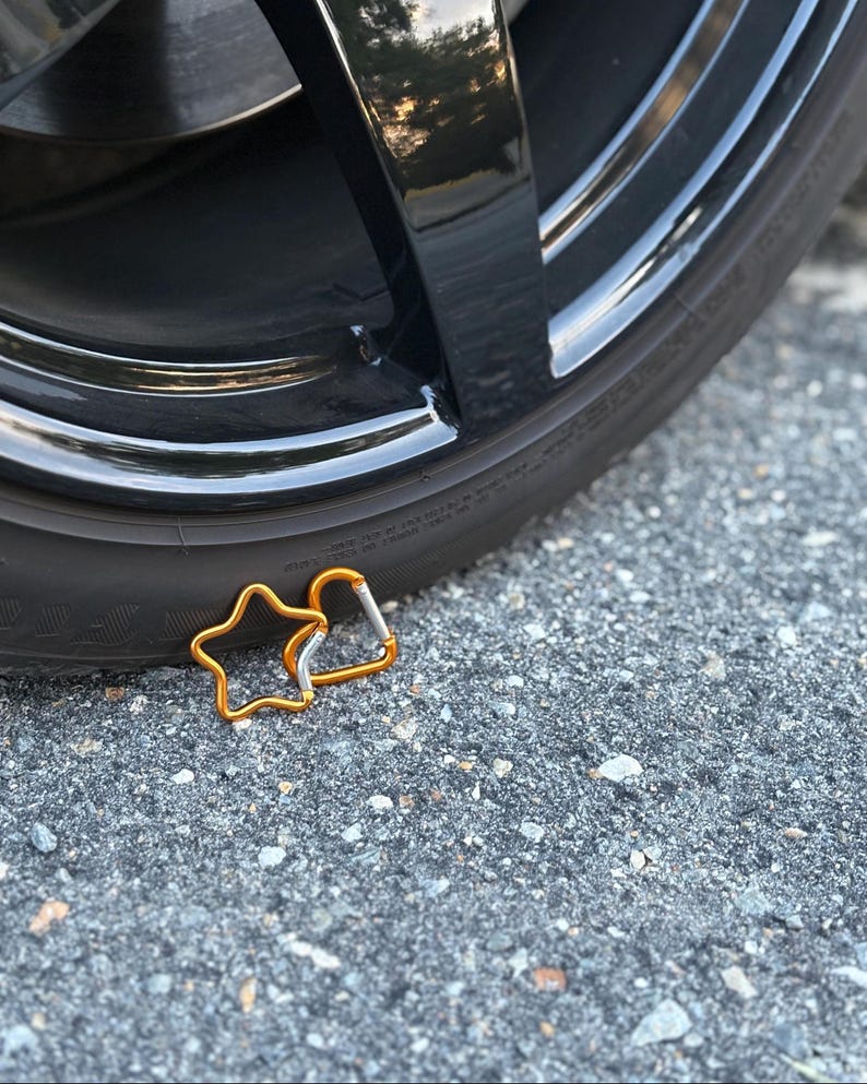 May include: Two gold-colored metal carabiners, one shaped like a five-pointed star and the other in a standard D-shape, featuring silver-toned spring-loaded gates, positioned near the base of a black rubber tire with a glossy rim.