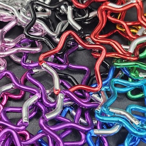Puede incluir: Una colecci&oacute;n de mosquetones con forma de estrella en varios colores met&aacute;licos, incluyendo rojo, azul, morado, verde, negro y plateado. Cada mosquet&oacute;n cuenta con un mecanismo de cierre de resorte que permite abrir y cerrar la estructura de estrella.