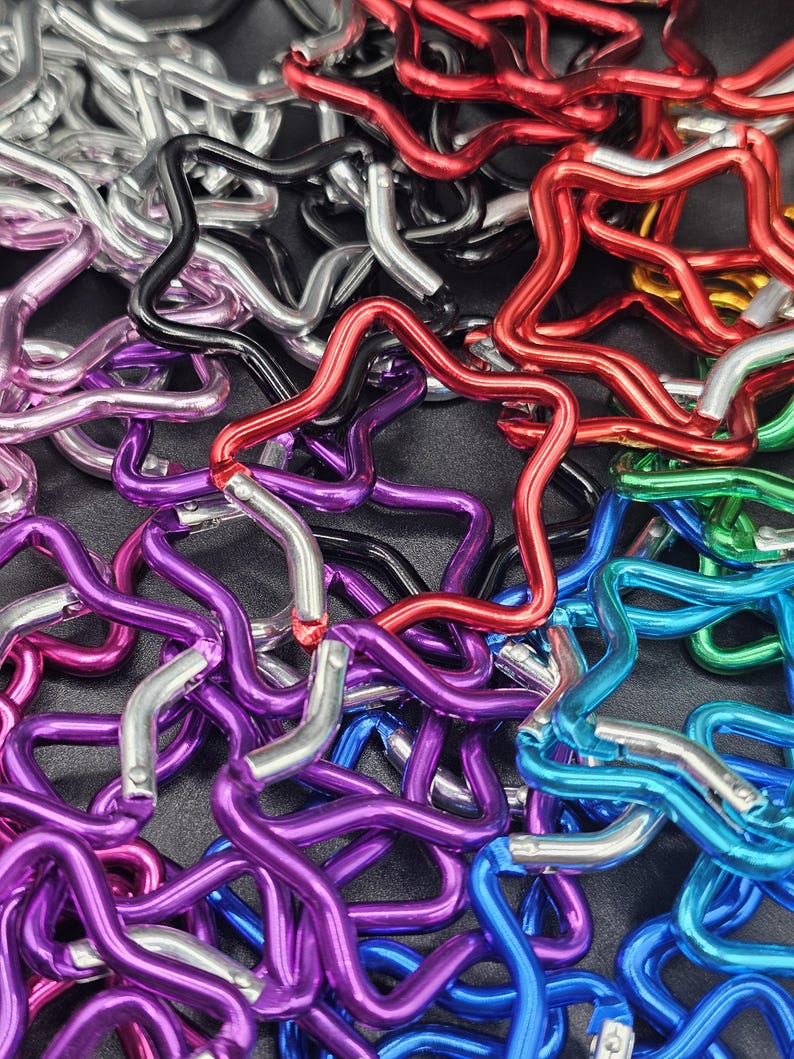 Puede incluir: Una colecci&oacute;n de mosquetones con forma de estrella en varios colores met&aacute;licos, incluyendo rojo, azul, morado, verde, negro y plateado. Cada mosquet&oacute;n cuenta con un mecanismo de cierre de resorte que permite abrir y cerrar la estructura de estrella.