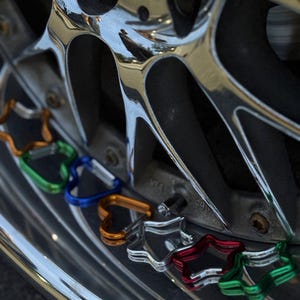 May include: A collection of small, heart-shaped metal carabiners in various colors including green, blue, orange, silver, and red, arranged along the curved, polished chrome rim of a vehicle wheel with visible spokes and a valve stem.