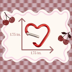 May include: A red heart-shaped carabiner with a silver-toned spring gate. The carabiner is displayed with measurement lines indicating a height and width of 1.75 inches. The design features a smooth, metallic finish and a functional opening mechanism.