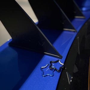 Puede incluir: Dos mosquetones en forma de estrella, uno de color azul met&aacute;lico y otro negro, descansan sobre una superficie azul brillante con estructuras de soporte verticales negras al fondo. Los mosquetones presentan un dise&ntilde;o de marco de alambre.
