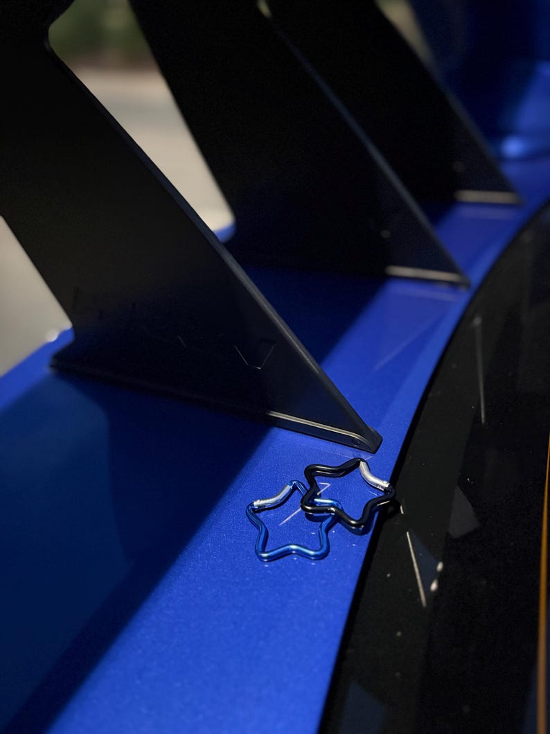 Puede incluir: Dos mosquetones en forma de estrella, uno de color azul met&aacute;lico y otro negro, descansan sobre una superficie azul brillante con estructuras de soporte verticales negras al fondo. Los mosquetones presentan un dise&ntilde;o de marco de alambre.