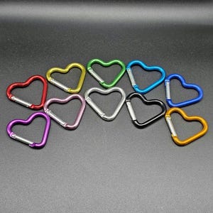 May include: A collection of eleven heart-shaped carabiners in various colors including red, yellow, green, light blue, dark blue, purple, pink, silver, black, and gold. Each carabiner features a spring-loaded metal gate for secure attachment.