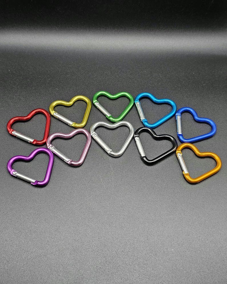 May include: A collection of eleven heart-shaped carabiners in various colors including red, yellow, green, light blue, dark blue, purple, pink, silver, black, and gold. Each carabiner features a spring-loaded metal gate for secure attachment.