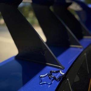 Puede incluir: Dos peque&ntilde;os mosquetones en forma de estrella, uno de color azul met&aacute;lico y otro negro, descansan sobre un aler&oacute;n de autom&oacute;vil azul brillante. El aler&oacute;n cuenta con soportes verticales negros y un acabado liso y brillante bajo luz natural.