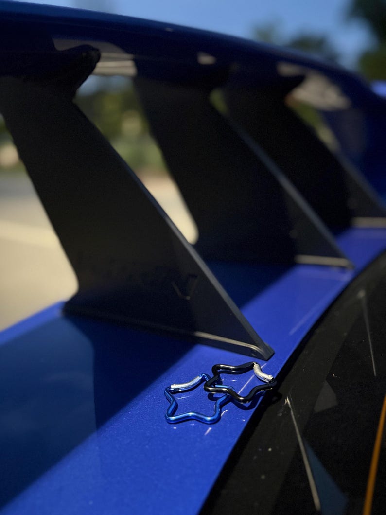 Puede incluir: Dos peque&ntilde;os mosquetones en forma de estrella, uno de color azul met&aacute;lico y otro negro, descansan sobre un aler&oacute;n de autom&oacute;vil azul brillante. El aler&oacute;n cuenta con soportes verticales negros y un acabado liso y brillante bajo luz natural.