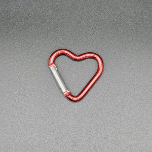 May include: A heart-shaped carabiner with a metallic red finish and a silver-colored spring-loaded gate mechanism. The carabiner features a smooth, rounded frame that forms a distinct heart shape, designed for securing items with a functional clip.