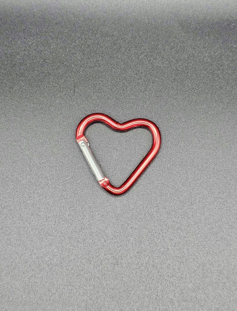 May include: A heart-shaped carabiner with a metallic red finish and a silver-colored spring-loaded gate mechanism. The carabiner features a smooth, rounded frame that forms a distinct heart shape, designed for securing items with a functional clip.