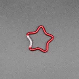 Puede incluir: Un mosquet&oacute;n con forma de estrella con un acabado rojo vibrante y un mecanismo de cierre de resorte de color plateado. El marco de metal forma el contorno de una estrella de cinco puntas, proporcionando un clip funcional para asegurar objetos.