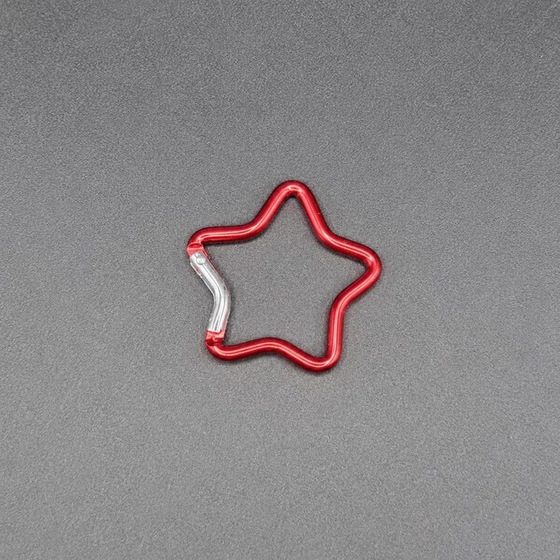 Puede incluir: Un mosquet&oacute;n con forma de estrella con un acabado rojo vibrante y un mecanismo de cierre de resorte de color plateado. El marco de metal forma el contorno de una estrella de cinco puntas, proporcionando un clip funcional para asegurar objetos.