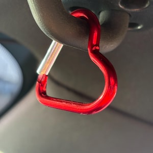 May include: A bright red heart-shaped carabiner clip with a metallic finish. The carabiner features a spring-loaded silver-toned gate mechanism on one side, allowing it to open and close securely while attached to a dark gray curved handle.