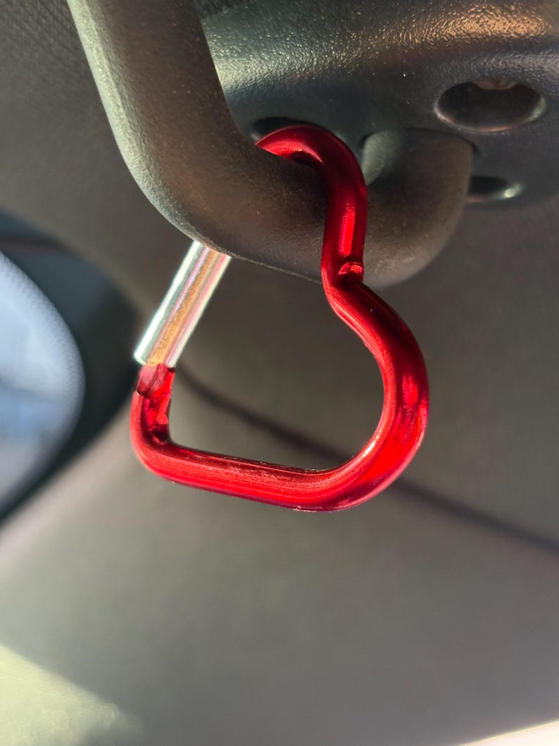 May include: A bright red heart-shaped carabiner clip with a metallic finish. The carabiner features a spring-loaded silver-toned gate mechanism on one side, allowing it to open and close securely while attached to a dark gray curved handle.