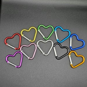 May include: A collection of ten heart-shaped carabiners in various colors including red, yellow, green, blue, purple, pink, silver, black, and orange. Each metal carabiner features a spring-loaded gate mechanism for secure attachment and closure.