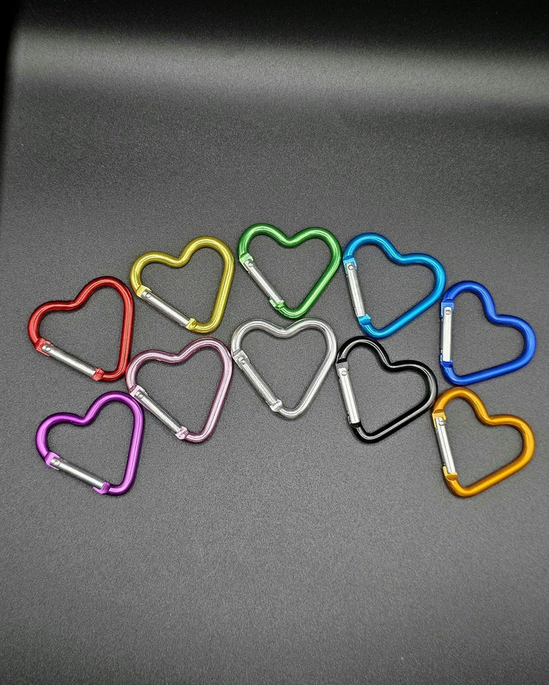 May include: A collection of ten heart-shaped carabiners in various colors including red, yellow, green, blue, purple, pink, silver, black, and orange. Each metal carabiner features a spring-loaded gate mechanism for secure attachment and closure.