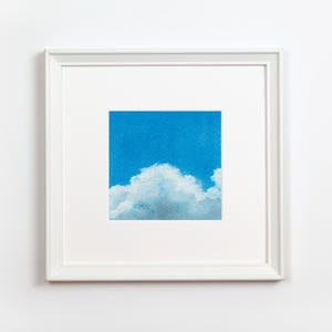 May include: A framed art print featuring a square image of a blue sky with fluffy white clouds. The artwork is encased in a white frame and mounted on a white wall, creating a clean and minimalist aesthetic.