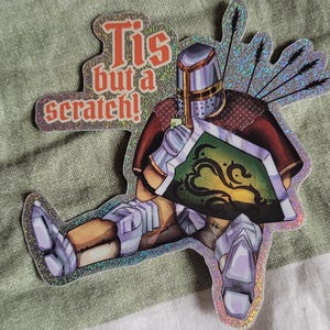 May include: A holographic sticker featuring a knight in silver armour and a brown tunic, holding a green shield with a black vine design. Several arrows are stuck in the knight's back. The text 'Tis but a scratch!' is printed in orange and red.