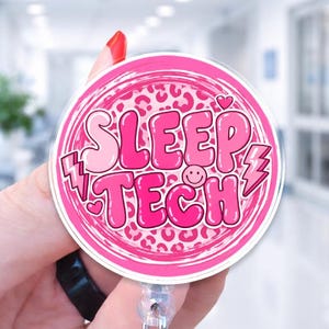 May include: A circular badge reel featuring a pink and white leopard print background with the text SLEEP TECH in bold, bubble-style pink lettering. The design includes small heart and lightning bolt accents, along with a small smiley face icon.