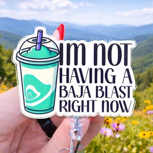 May include: A badge reel featuring a graphic of a teal slushy drink in a clear plastic cup with a purple straw, accompanied by the black text "I'M NOT HAVING A BAJA BLAST RIGHT NOW" on a white background with a clear acrylic border.