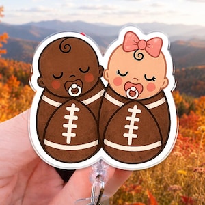 May include: An acrylic badge reel featuring two cartoon babies wrapped in brown football-patterned swaddles. Both babies have closed eyes, pacifiers, and dark brown football-shaped bodies with white laces. One baby has a pink bow on its head.