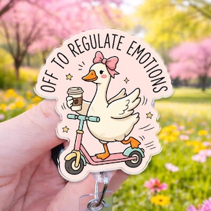 May include: A clear acrylic badge reel featuring a cartoon goose with a pink bow, riding a pink scooter while holding a coffee cup. The text OFF TO REGULATE EMOTIONS is printed in black around the top of the goose, surrounded by small yellow stars.