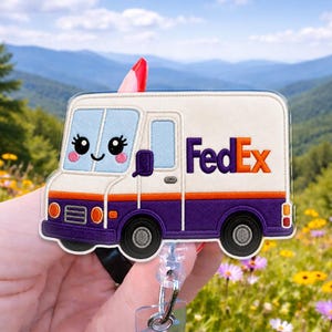 May include: A badge reel featuring a cream and purple embroidered FedEx delivery truck with a smiling face, large eyes, and pink cheeks. The truck has a clear acrylic overlay and is attached to a retractable cord mechanism for holding identification.