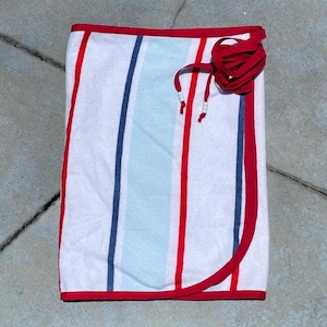 May include: A folded beach towel with vertical stripes in red, blue, and light blue on a white background. The towel is trimmed with red edging and has a red tie closure. The towel is made of a soft, absorbent material.