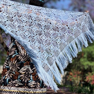 May include: A light blue crochet shawl with fringe. The shawl is draped over a person wearing a patterned dress.