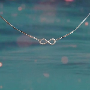 May include: A delicate silver necklace featuring an infinity symbol pendant. The pendant is centered on a fine chain, set against a blurred teal background with soft, out-of-focus highlights. The necklace is a simple, elegant piece of jewelry.