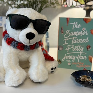 May include: A white polar bear plush toy wearing black sunglasses and a red and gray scarf. The bear is sitting on a white surface next to a teal book titled "The Summer I Turned Pretty" by Jenny Han. A small blue dish with a gold seashell is also on the surface.