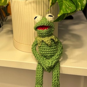 May include: A handmade green crochet frog doll with white eyes and a red mouth. The frog is sitting with its arms crossed. The doll is a whimsical decorative item.