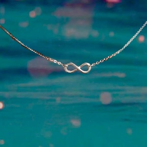 May include: A delicate silver necklace featuring an infinity symbol pendant. The pendant is the focal point, suspended on a fine chain. The background is a blurred teal color with soft, out-of-focus highlights, creating a dreamy aesthetic.