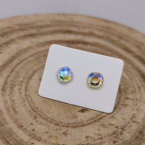 May include: Two iridescent square-cut crystal stud earrings on a white card. The crystals reflect a spectrum of colors, including blues, greens, and yellows. The earrings are displayed on a wooden surface.